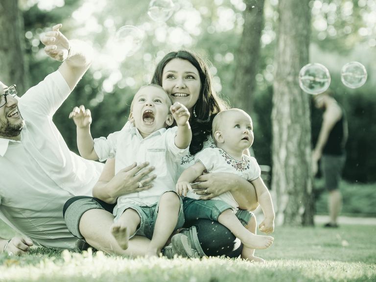 A laughing family of four on a picnic blanket in a sunny park, the father lifting their toddler